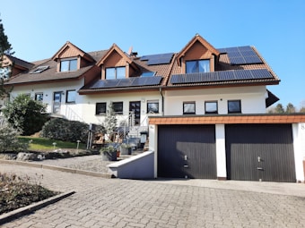 A row of modern, suburban houses with sloping roofs, featuring solar panels installed on the roof of the central house. The exteriors are painted white, complemented by brown roofs and windows. A neatly paved driveway and a garden with shrubs and small trees are visible in the foreground, leading up to the garages of the central house.