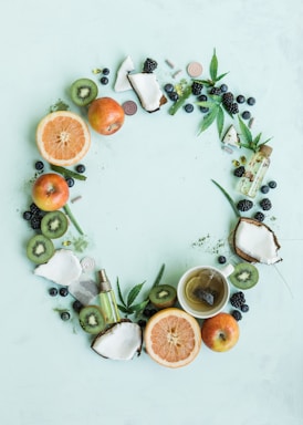 A circular arrangement of fresh fruits and natural elements including apples, kiwis, grapefruit halves, coconuts, leaves, and blackberries. There is also a tea cup filled with green tea and a dropper bottle. The items are placed on a light mint background, giving a fresh and natural look.