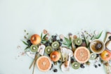 A variety of fresh fruits including apples, grapefruit, kiwis, and blackberries are arranged alongside loose tea leaves, makeup brushes, and bottles of skincare products on a light green background. The composition is artistic and balanced, incorporating natural elements with beauty products.