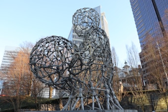A large abstract sculpture composed of interconnected figures is set against a backdrop of tall modern buildings and bare trees. The sculpture appears to be made of metal and features a series of human-like forms creating a cohesive structure. The clear sky and cityscape provide a contrasting modern urban environment.