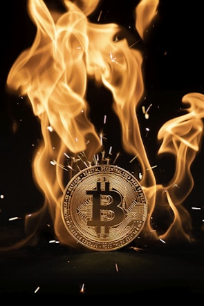 A Bitcoin coin engulfed in bright orange and yellow flames with sparks flying off. The fire creates a striking contrast against the dark background.