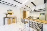 A modern, minimalist kitchen area with a light color palette. Features include a wooden breakfast bar with two woven chairs, a compact kitchen setup with shelves holding dishes and kitchenware, and a small fridge. On the adjacent wall, there's a decorative console table with two large baskets underneath, a circular mirror above, and a potted plant along with a few decorative items.
