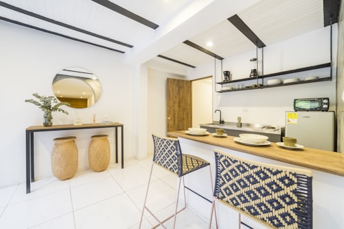 A modern, minimalist kitchen area with a light color palette. Features include a wooden breakfast bar with two woven chairs, a compact kitchen setup with shelves holding dishes and kitchenware, and a small fridge. On the adjacent wall, there's a decorative console table with two large baskets underneath, a circular mirror above, and a potted plant along with a few decorative items.