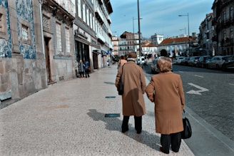A sophisticated couple dressed in chocolate brown and beige vintage-inspired clothing, walking hand in hand along an Italian villa terrace.