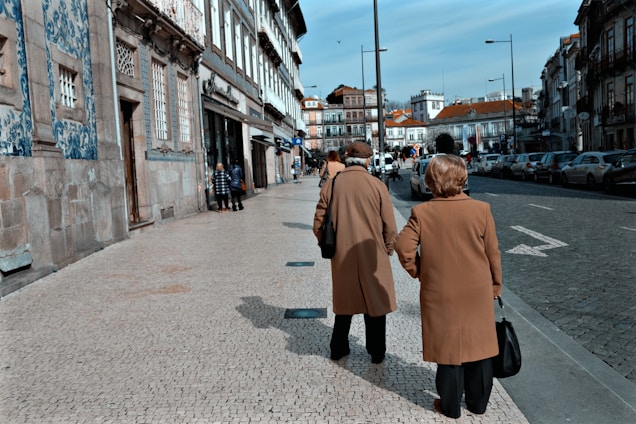 A sophisticated couple dressed in chocolate brown and beige vintage-inspired clothing, walking hand in hand along an Italian villa terrace.