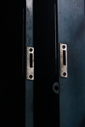 Two metal doors with a dark, weathered texture are slightly ajar, revealing a narrow gap of darkness between them. The doors have metallic slots and screws, suggesting they may belong to a cabinet or a locker.