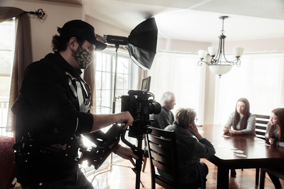 A camera operator wearing a harness and a camouflage mask is filming a group of four people sitting around a dining table. The setting is in a bright room with large windows and overhead chandelier. One person is operating a camera with a large attached light source, focusing on the seated group.