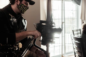 A person is operating a professional camera with a stabilizing rig. They are wearing a camouflage face mask and a black outfit. The background shows a bright window with curtains and a partial view of a chair.