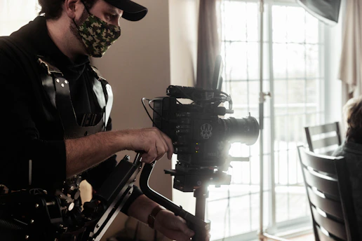 A person is operating a professional camera with a stabilizing rig. They are wearing a camouflage face mask and a black outfit. The background shows a bright window with curtains and a partial view of a chair.