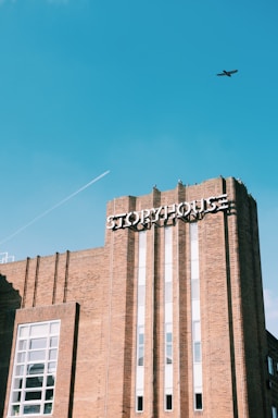 A brick building with the word 'Storyhouse' prominently displayed on the facade. The architecture features vertical lines and large windows. In the clear blue sky above, there is a small airplane and a contrail.