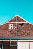 A brick building with a gable roof and a sky-blue background. The building has a large 'R2' painted in white on the brickwork near the peak of the roof. There is a black light fixture attached to the wall below the 'R2' sign, and white trim outlines the gable. Part of a window frame is visible at the bottom of the image.