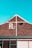A brick building with a gable roof and a sky-blue background. The building has a large 'R2' painted in white on the brickwork near the peak of the roof. There is a black light fixture attached to the wall below the 'R2' sign, and white trim outlines the gable. Part of a window frame is visible at the bottom of the image.
