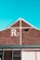 A brick building with a gable roof and a sky-blue background. The building has a large 'R2' painted in white on the brickwork near the peak of the roof. There is a black light fixture attached to the wall below the 'R2' sign, and white trim outlines the gable. Part of a window frame is visible at the bottom of the image.