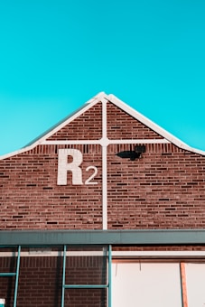 A brick building with a gable roof and a sky-blue background. The building has a large 'R2' painted in white on the brickwork near the peak of the roof. There is a black light fixture attached to the wall below the 'R2' sign, and white trim outlines the gable. Part of a window frame is visible at the bottom of the image.