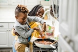 A young boy is pouring sauce into a skillet on the stove with the help of a smiling woman standing behind him. The scene is set in a kitchen, with a modern stove and white cabinetry. The countertop holds ingredients like fresh vegetables, a bag of wholesome meats, and a spoon resting on the skillet.