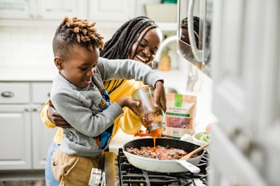 A young boy is pouring sauce into a skillet on the stove with the help of a smiling woman standing behind him. The scene is set in a kitchen, with a modern stove and white cabinetry. The countertop holds ingredients like fresh vegetables, a bag of wholesome meats, and a spoon resting on the skillet.