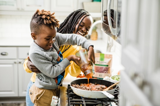 A young boy is pouring sauce into a skillet on the stove with the help of a smiling woman standing behind him. The scene is set in a kitchen, with a modern stove and white cabinetry. The countertop holds ingredients like fresh vegetables, a bag of wholesome meats, and a spoon resting on the skillet.