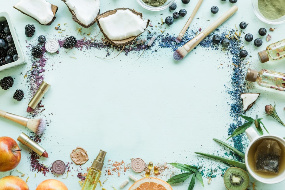 A flat lay arrangement features various natural and cosmetic items on a light green surface. Surrounding an empty center are brushes, sliced fruits like apples and kiwi, makeup items such as lipsticks and foundation, loose tea leaves, berries like blueberries and blackberries, and cut coconut. There are also small bottles containing oil and scattered, colorful powders.