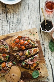 Slices of gourmet pizza topped with cherry tomatoes, zucchini, and fresh basil rest on a rustic wooden cutting board. A pizza cutter lies nearby, alongside a small white dish containing dark sauce and a glass of pink beverage. The surface is textured with a weathered wooden appearance.