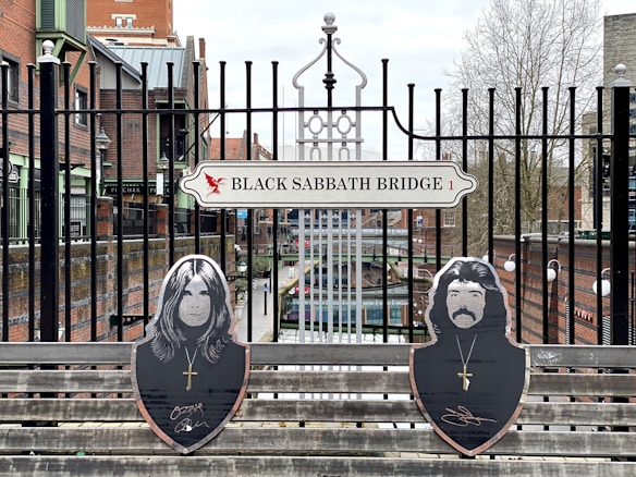 A sign reading 'Black Sabbath Bridge 1' is mounted on a metal fence. Below it, two wooden cutouts of faces with long hair and necklaces are attached to a bench. The background features a canal, buildings, and bare trees under a cloudy sky.