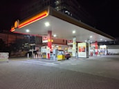 A well-lit gas station at night featuring multiple fuel pumps under a large canopy with a prominent Shell logo. The area is organized, with various signage for fueling options, including Shell V-Power. A car is parked near the pump, and the perimeter is marked with orange traffic cones. Bright lighting from above highlights the canopy and the pump area in contrast to the dark surroundings.
