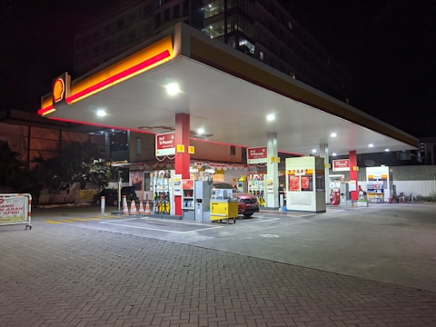 A well-lit gas station at night featuring multiple fuel pumps under a large canopy with a prominent Shell logo. The area is organized, with various signage for fueling options, including Shell V-Power. A car is parked near the pump, and the perimeter is marked with orange traffic cones. Bright lighting from above highlights the canopy and the pump area in contrast to the dark surroundings.