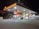 A well-lit gas station at night featuring multiple fuel pumps under a large canopy with a prominent Shell logo. The area is organized, with various signage for fueling options, including Shell V-Power. A car is parked near the pump, and the perimeter is marked with orange traffic cones. Bright lighting from above highlights the canopy and the pump area in contrast to the dark surroundings.