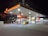 A well-lit gas station at night featuring multiple fuel pumps under a large canopy with a prominent Shell logo. The area is organized, with various signage for fueling options, including Shell V-Power. A car is parked near the pump, and the perimeter is marked with orange traffic cones. Bright lighting from above highlights the canopy and the pump area in contrast to the dark surroundings.