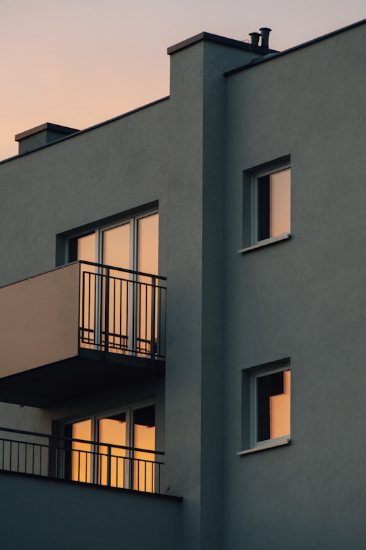 A modern apartment building with a minimalist design, featuring balconies and multiple windows reflecting a warm sunset light. The walls are painted in a muted gray color, and the scene is captured at dusk with a soft, ambient glow.