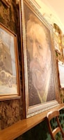A gallery wall displays a collection of framed art pieces. The most prominent piece is a large portrait in the center, surrounded by gold moldings, featuring a man with a distinct expression and vivid brush strokes. Other paintings, including a rustic scene with a figure and a snow-covered landscape, hang around it. The wall is decorated with lush greenery, adding a natural texture to the artistic arrangement. A wooden chair is positioned in front of the paintings.