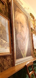 A gallery wall displays a collection of framed art pieces. The most prominent piece is a large portrait in the center, surrounded by gold moldings, featuring a man with a distinct expression and vivid brush strokes. Other paintings, including a rustic scene with a figure and a snow-covered landscape, hang around it. The wall is decorated with lush greenery, adding a natural texture to the artistic arrangement. A wooden chair is positioned in front of the paintings.