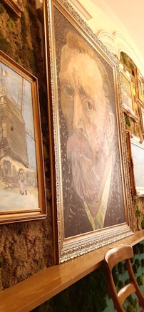 A gallery wall displays a collection of framed art pieces. The most prominent piece is a large portrait in the center, surrounded by gold moldings, featuring a man with a distinct expression and vivid brush strokes. Other paintings, including a rustic scene with a figure and a snow-covered landscape, hang around it. The wall is decorated with lush greenery, adding a natural texture to the artistic arrangement. A wooden chair is positioned in front of the paintings.