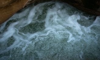 Foamy water swirls over a rocky surface, creating a textured pattern of bubbles and white foam against a dark green background. The image captures the dynamic movement of water as it interacts with the rock.