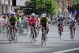 Cyclists are participating in a road race or cycling event in an urban setting, surrounded by onlookers and safety barriers. Riders are wearing helmets and cycling jerseys, and they appear focused and determined while pedaling down the road.