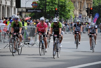 Cyclists are participating in a road race or cycling event in an urban setting, surrounded by onlookers and safety barriers. Riders are wearing helmets and cycling jerseys, and they appear focused and determined while pedaling down the road.