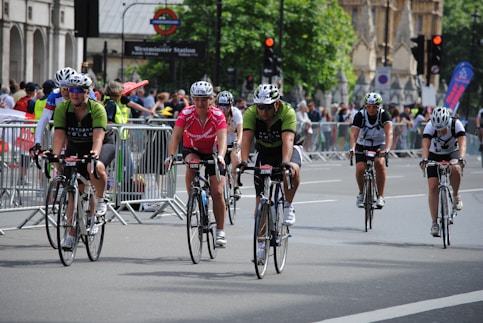 Cyclists are participating in a road race or cycling event in an urban setting, surrounded by onlookers and safety barriers. Riders are wearing helmets and cycling jerseys, and they appear focused and determined while pedaling down the road.