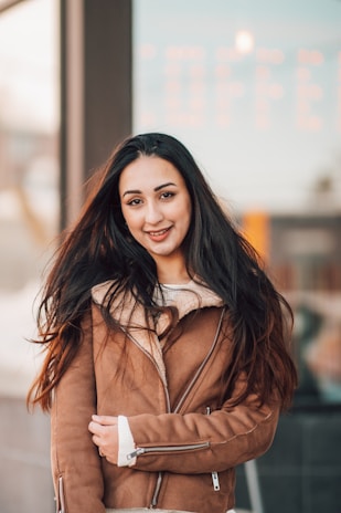 A woman with long dark hair is wearing a brown shearling jacket and a white sweater. She is outdoors with a blurred background that suggests a cityscape or urban setting during the daytime.