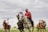 A group of people are playing polo on a grassy field. They are riding horses and holding mallets, wearing helmets for protection. The sky is overcast, creating a subdued atmosphere.