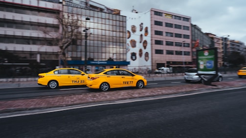 A pair of yellow taxis driving along a city street with buildings in the background. The taxis have black lettering on the sides and are moving past modern office buildings. The street is lined with trees and other vehicles are visible in the distance.