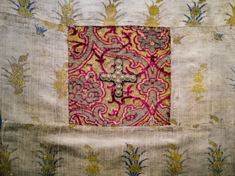 A detailed textile with an ornate design. The center features a square section with a cross decorated with small pearls and surrounded by intricate, swirling patterns in gold, pink, and green. The surrounding fabric is plain with embroidered golden and blue motifs.