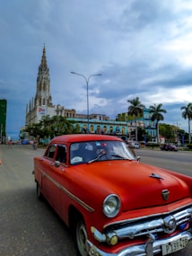 A vintage red car is parked on a wide street with an old cathedral in the background. Palm trees stand along the roadside, and colorful colonial-style buildings are visible. The sky is overcast, suggesting a cloudy or rainy day.