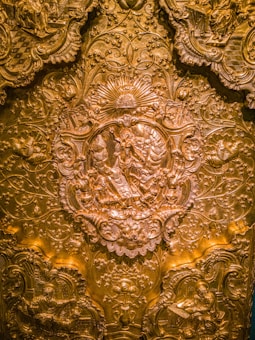 Intricate gold relief work featuring detailed carvings of religious imagery. Central figures are surrounded by ornate patterns, including floral motifs and decorative scrollwork. The craftsmanship is highly detailed, giving the piece a rich and luxurious appearance.