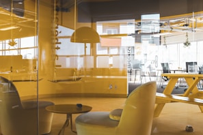 An office space with modern furniture including yellow chairs, a table, a large communal table with benches, and hanging lamps. The room is enclosed by glass walls. Natural light enters through large windows, creating a bright atmosphere. Some plants hang near the ceiling, adding a touch of greenery.