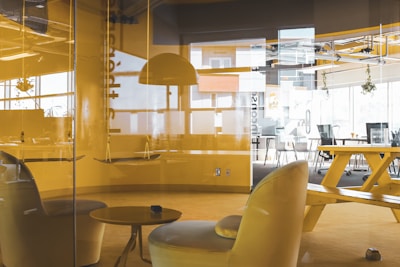 An office space with modern furniture including yellow chairs, a table, a large communal table with benches, and hanging lamps. The room is enclosed by glass walls. Natural light enters through large windows, creating a bright atmosphere. Some plants hang near the ceiling, adding a touch of greenery.
