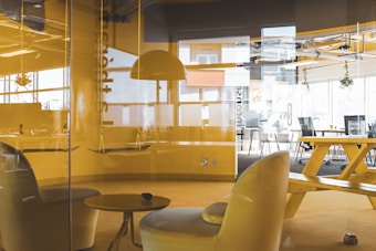 An office space with modern furniture including yellow chairs, a table, a large communal table with benches, and hanging lamps. The room is enclosed by glass walls. Natural light enters through large windows, creating a bright atmosphere. Some plants hang near the ceiling, adding a touch of greenery.