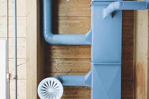 A section of industrial ductwork is mounted on a wooden ceiling, featuring a large blue metal air duct and a circular air vent. The ductwork components are prominently visible against a backdrop of wooden panels with a light finish.