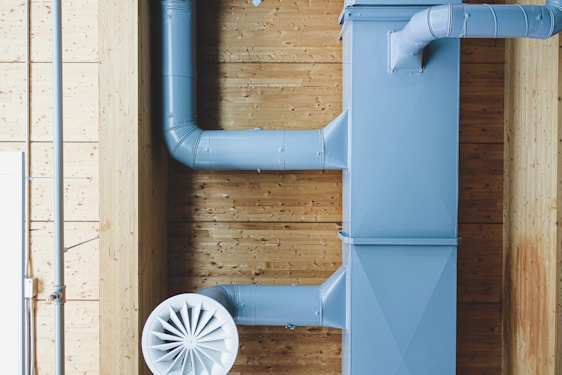 A section of industrial ductwork is mounted on a wooden ceiling, featuring a large blue metal air duct and a circular air vent. The ductwork components are prominently visible against a backdrop of wooden panels with a light finish.