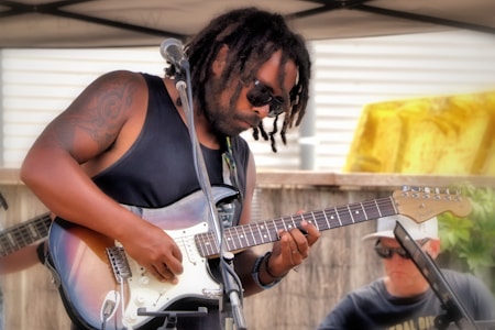 A man with sunglasses and dreadlocks plays an electric guitar under a canopy. He is wearing a sleeveless black shirt, showcasing a tattoo on his right arm. Another person, also wearing sunglasses and a cap, can be seen in the background.