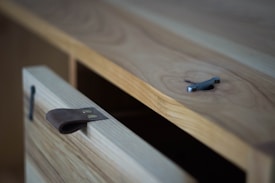 A close-up view of a wooden cabinet with a leather strap handle. The cabinet door is slightly open, showing the craftsmanship and natural wood grain. A metal object is placed on top of the cabinet.