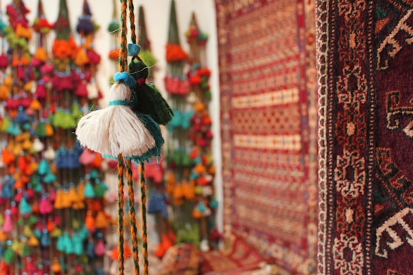 A collection of colorful tassels hanging on a wall is complemented by intricately patterned rugs with rich, warm hues. The focus is on the textures and vibrant colors, creating an inviting and artistic atmosphere.